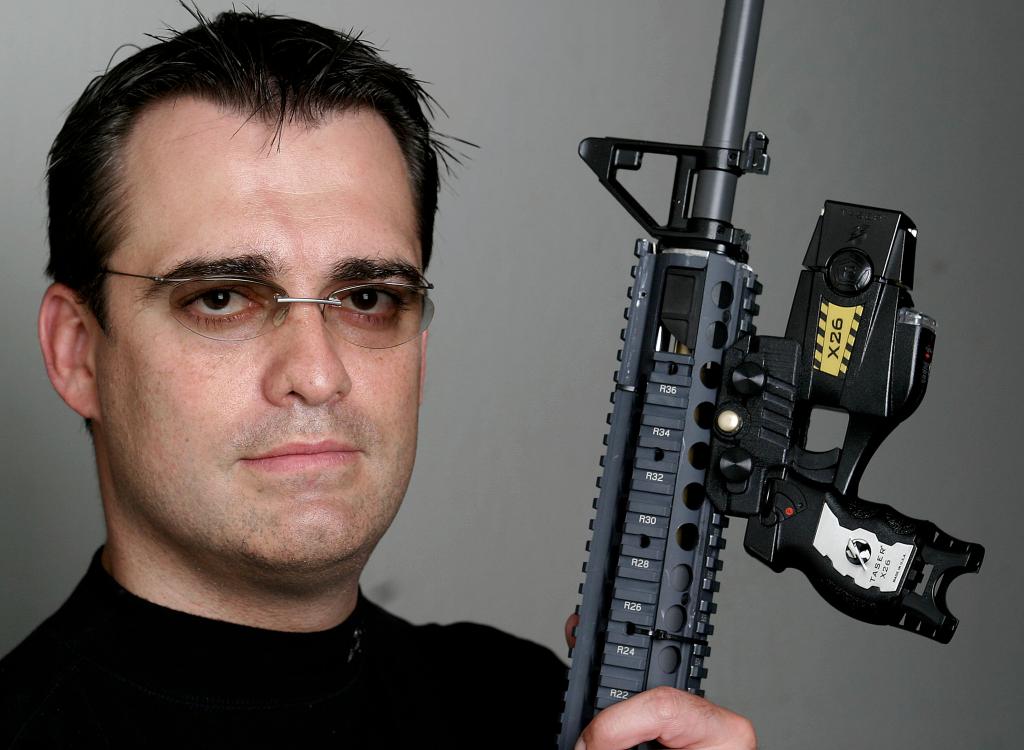 A man holding a rifle with a Taser X26 attached.