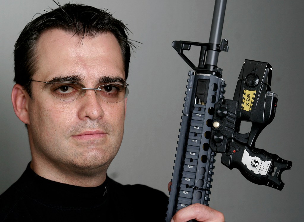 A man holding a rifle with a Taser X26 attached.